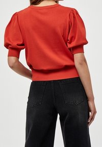 Punainen hihansuista poimutettu neule, jossa on resorireunus hihansuissa ja helmassa, yhdistetty mustiin, korkeavyötäröisiin farkkuihin, joissa on takataskut. Sileä kangaspinta.