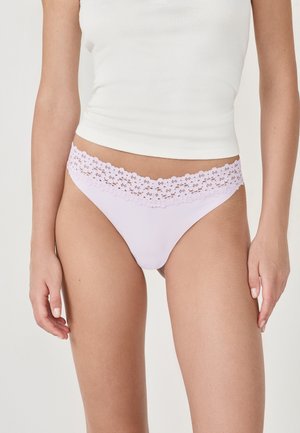Donna che indossa una lingerie bikini in pizzo lavanda e un top senza maniche bianco, in piedi contro uno sfondo chiaro e uniforme.