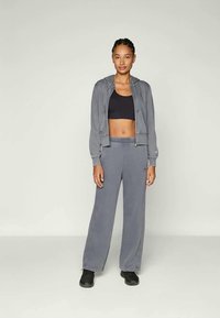 Mujer joven de pie, vestida con una sudadera gris con cremallera, pantalones de chándal a juego, sujetador deportivo negro y zapatillas negras sobre un fondo neutro.