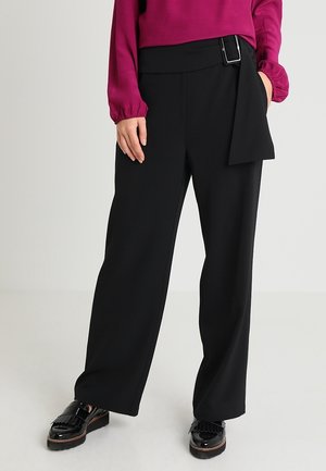 Osoba ubrana w czarne szerokie spodnie z klamrą, magentową bluzkę z długim rękawem oraz czarne błyszczące mokasyny, stojąca na jasnej podłodze.