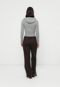 Persona de pie de espaldas a la cámara, vistiendo un suéter con capucha gris ajustado, pantalones oscuros marrón y zapatos burdeos sobre un fondo blanco.