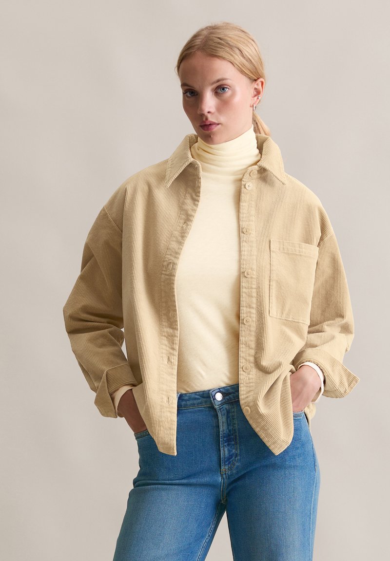 Beżowa koszula sztruksowa z guzikiem z przodu, dużym kołnierzem i kieszenią na piersi, noszona na jasnym golfie i połączona z niebieskimi dżinsami.