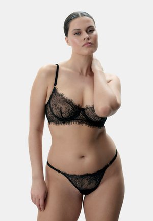 Vrouw met naar achteren gekamd haar die een zwart kanten bh en bijpassend ondergoed draagt, poseert met één hand aan haar nek tegen een effen achtergrond.