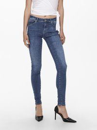 Jeans de ganga azuis skinny com uma cintura média, bolsos frontais e um leve desbotado. Combinados com saltos altos pretos pontiagudos e uma blusa branca envolvente.