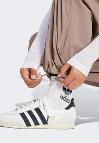 Vita Adidas-sneakers med svarta ränder, beige träningsbyxor och en vit långärmad tröja. Händer drar i snöret från byxorna.