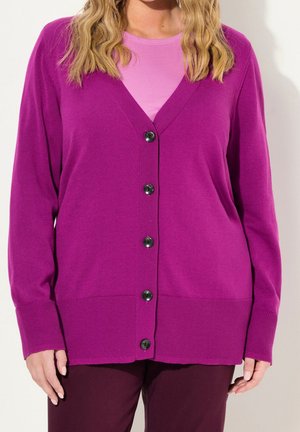 Kvinde iført en knaldfarvet lilla knapcardigan over en pink top og mørkelilla bukser, med løst bølget blondt hår synligt fra nakken og ned.