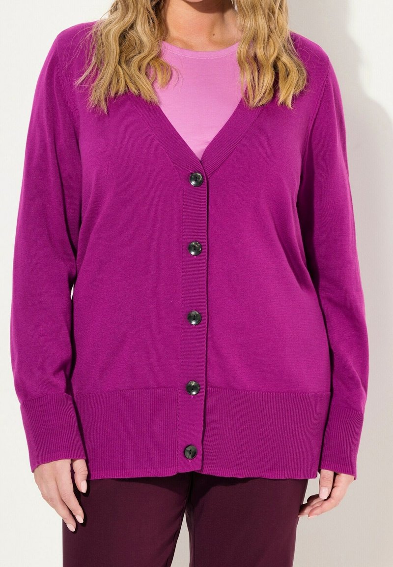 Femme portant un cardigan violet vif boutonné sur un haut rose et un pantalon violet foncé, avec des cheveux blonds ondulés et lâches visibles du cou vers le bas.