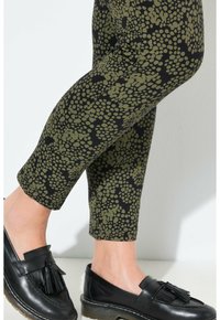 Gambe che indossano pantaloni neri tagliati con un motivo astratto a pois verde oliva e mocassini neri in pelle con nappine su sfondo chiaro.