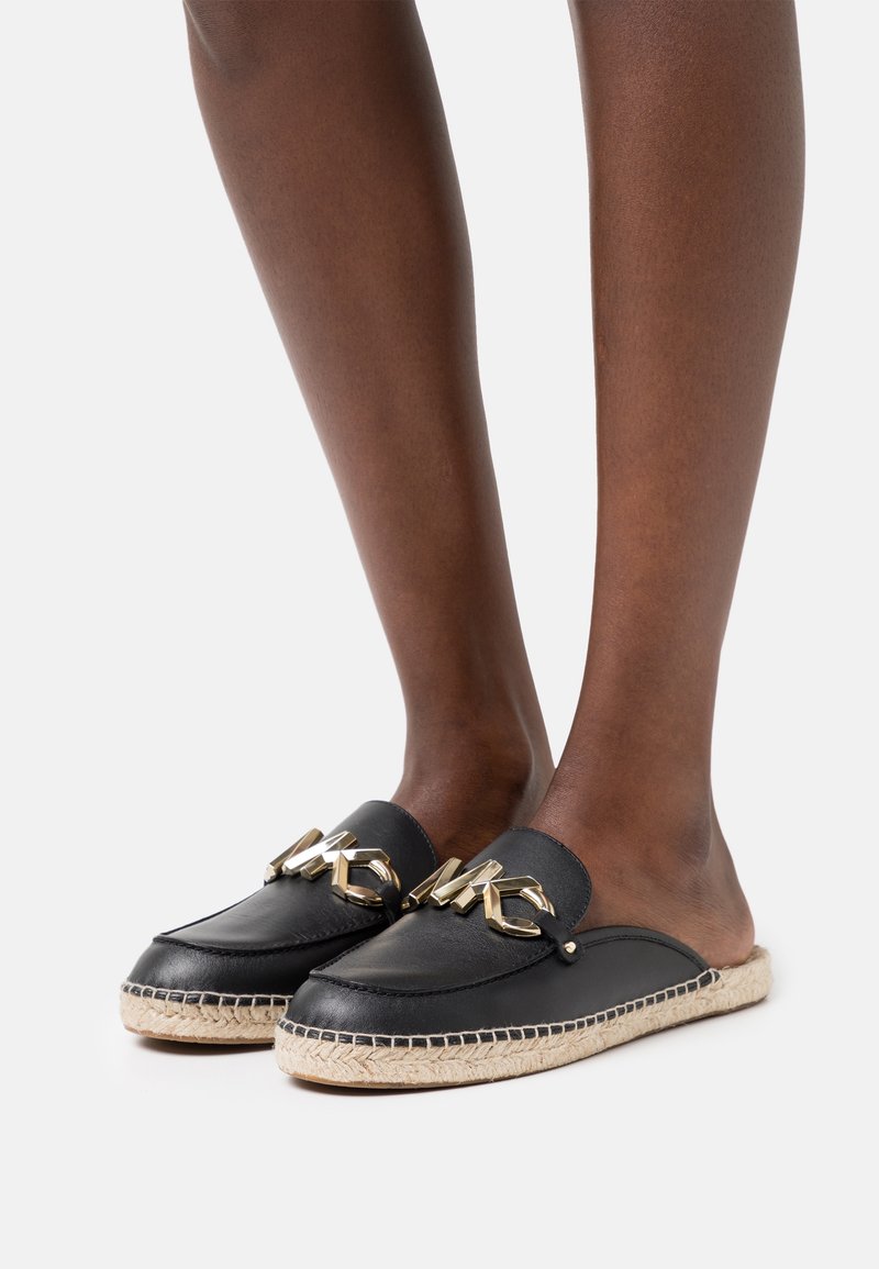 Pieds chaussés de mocassins espadrilles noirs en cuir à enfiler, ornés d'un logo MK doré et de semelles en jute tressé, sur fond blanc.
