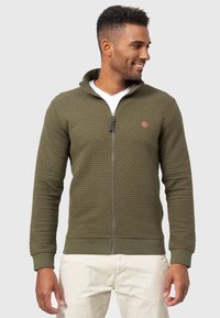 Chaqueta de color verde oliva con cremallera, fabricada en un tejido texturizado, cuello alto, puños acanalados y logotipo bordado. Llevada sobre una camiseta blanca.