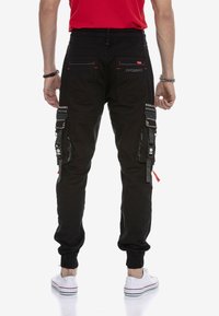 Schwarze Cargo-Hosen mit elastischen Bündchen, ausgestattet mit mehreren Seitentaschen, roten Akzenten und einem strukturierten Material. Branding auf der Rückseite.