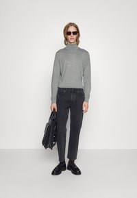 Grå rullekragegenser, sorte smale jeans, sorte dressko, holder en svart veske, solbriller med rektangulær ramme, nøytral bakgrunn.