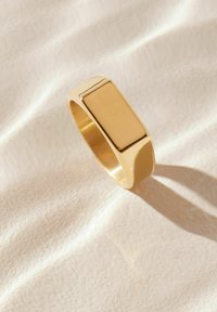Goldfarbener Ring mit flacher rechteckiger Front, glatter Oberfläche und rundem Band, der einen subtilen Schatten auf einer hellen Stoffoberfläche wirft.