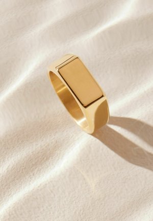 Inel cu ton auriu, cu o față rectangulară plată, finisaj neted și o bandă rotunjită, care proiectează o umbră subtilă pe o suprafață de țesătură deschisă.