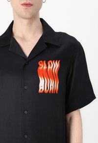 Korte mouwen zwarte shirt gemaakt van een gladde stof met een verloop oranje grafische tekst "SLOW BURN" op de linkerkant van de borst.