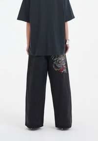 Weite schwarze Jeans mit einem bunten gestickten Drachendesign auf der linken Seite, kombiniert mit einem locker sitzenden dunkelgrauen T-Shirt.