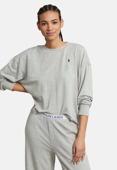 Νεαρή γυναίκα με πλεξούδες, ντυμένη με γκρι μακρυμάνικο φούτερ και αντίστοιχο παντελόνι φόρμας με ορατή μέση Ralph Lauren.