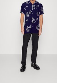 Tmavě modrá košile s krátkým rukávem s bílým paisley vzorem, límcem a knoflíky, doplněná černými kalhotami a lesklými černými botami.