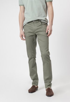 Hombre vistiendo pantalones rectos de color verde oliva, camiseta polo de manga corta a rayas y mocasines de ante marrones, de pie frente a un fondo liso.