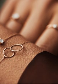 Roze gouden armband met twee in elkaar grijpende cirkels; delicate ketting; gladde textuur; gedragen op een gestructureerde huidoppervlak met ringen op de achtergrond.