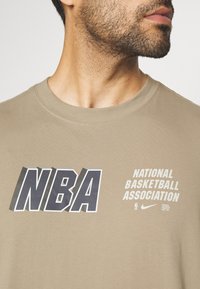 Camiseta color caqui con un audaz gráfico de "NBA" en azul oscuro y el texto "ASOCIACIÓN NACIONAL DE BALONCESTO" en blanco. Material de algodón suave.