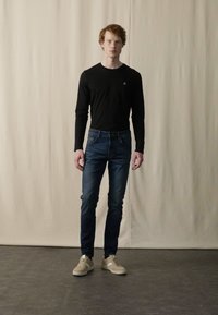 Chemise noire à manches longues, jean slim-fit bleu et baskets beiges, sur un fond neutre, avec un détail de logo subtil sur la chemise.