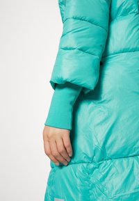 Main reposant à côté d'une manche de doudoune turquoise dépassant d'un poignet côtelé assorti sur un fond blanc.