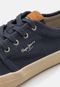 Baskets en toile bleu marine avec semelle en caoutchouc blanche. Présentent des œillets en métal, des lacets texturés, et un accent en cuir marron sur le talon avec détail de logo.