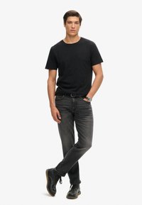 Zwarte katoenen T-shirt, gecombineerd met donkergrijze spijkerbroek. Het model draagt zwarte lederen schoenen met een lichtbruine zool, met minimale details.