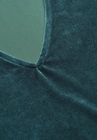 Teal sammetstyg med en slät textur, med en djup v-ringning och en kontrasterande grön innerfoder. Diskret samlingsdetalj ingår.