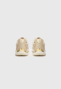 Scarpe da ginnastica beige con una tomaia testurizzata e scintillante, dettagli dorati sul tallone e una suola bianca imbottita. Viste da dietro.