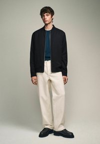 Chaqueta bomber negra, camisa azul verdosa, pantalones de pierna ancha color crema y zapatos negros con suela texturizada, contra un fondo neutral.