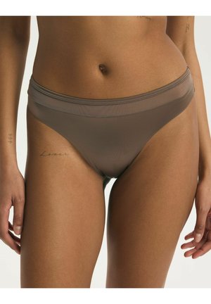 Žena nosiace taupe bezšvové nohavičky s vysokým vystrihnutím, ukazujúca tetovania na ruke a stehne, na neutrálnom pozadí.