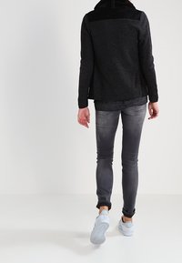 Osoba odchodząca w ciemnoszarych dżinsach, czarnym swetrze i białych sneakersach na jednolitym białym tle.