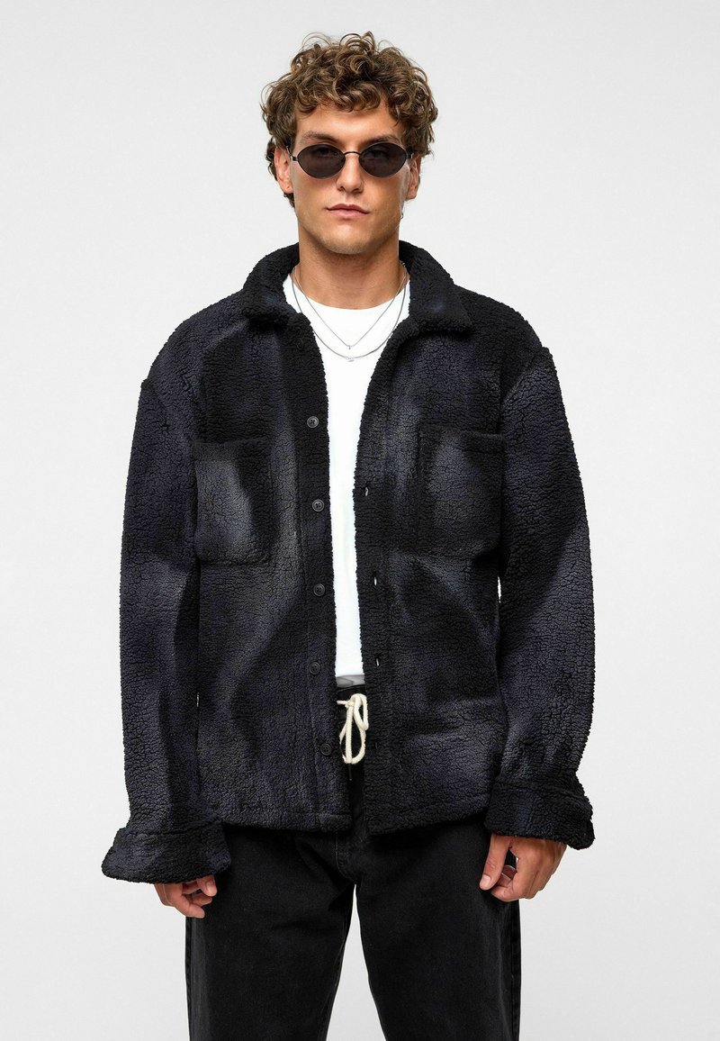 Zwarte, fuzzy fleece shirtjack met twee voorzakken, knoopsluiting en omgeslagen kraag. Geko zen over een wit T-shirt, gecombineerd met zwarte broek.