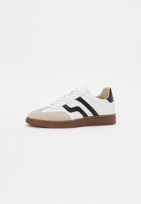 White sneaker with black stripes, suede beige toe cap, and brown rubber sole. Lace-up design, padded collar, and textured surface.