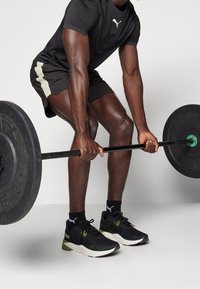 Sort atletisk skjorte og shorts med beige accenter; løfter en vægtstang; iført sorte og grønne sneakers; stærke muskuløse ben.