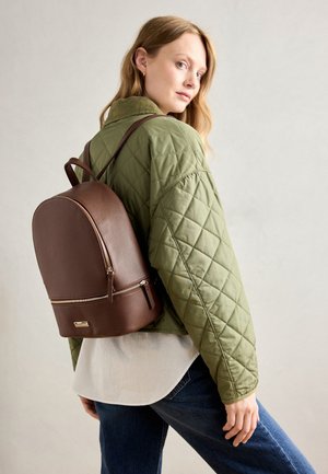 Kvinde i grøn quiltet jakke og jeans iført en brun læderrygsæk, der ser over skulderen mod en neutral baggrund.