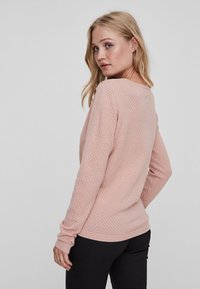 Lys rosa teksturert genser med honningkake-mønster, lange ermer og rund hals. Ribbestrikkede mansjetter og nederkant. Svarte bukser synlige.