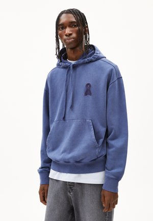 Blaue Baumwollkapuzenjacke mit einer Kängurutasche, einer verstellbaren Kapuze und einem strukturierten Logo-Akzent auf der Brust. Über einem weißen Hemd getragen.