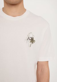 Camiseta blanca de algodón con mangas cortas que presenta un pequeño diseño bordado de palmeras y "AX" en tonos verdes y dorados.