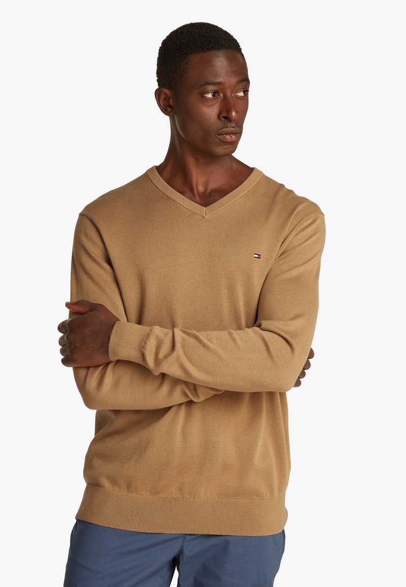 Homme aux cheveux courts portant un pull beige à col en V et un pantalon bleu, bras croisés, regardant vers la droite sur un fond blanc uni.