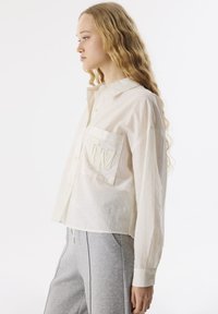 Chemise blanche à manches longues en tissu léger, avec un col pointu et une poche sur la poitrine gauche avec un design embossé.
