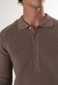 Pull en maille côtelée marron avec un col et un plastron à cinq boutons. Présente une texture douce et un design classique.