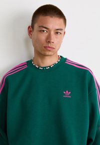 Green sweatshirt with pink stripes on shoulders, featuring an embroidered pink Adidas logo on the chest, worn with a beaded necklace.