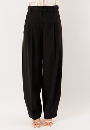 Pantaloni neri a vita alta con gamba larga, pieghe frontali, chiusura con doppio bottone e spacchi laterali, indossati con sandali color crema.