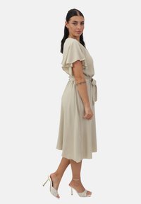 Vestido beige hasta la rodilla con mangas acampanadas, cintura ceñida y textura suave; combinado con sandalias de tacón blanco con un patrón trenzado.