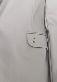 Šedá obleková sako s hladkým povrchem, klopovým límcem a stříbrným monogramem na kapsičce na rukávu.