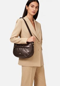 Borsa a tracolla in pelle metallizzata marrone con forma curva, tasca frontale con zip e tracolla in rilievo. Presenta una lieve lucentezza.