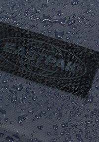 Tela de color azul marino oscuro con gotas de agua, con un parche del logo negro de Eastpak. Se puede apreciar la superficie texturizada y el material duradero.